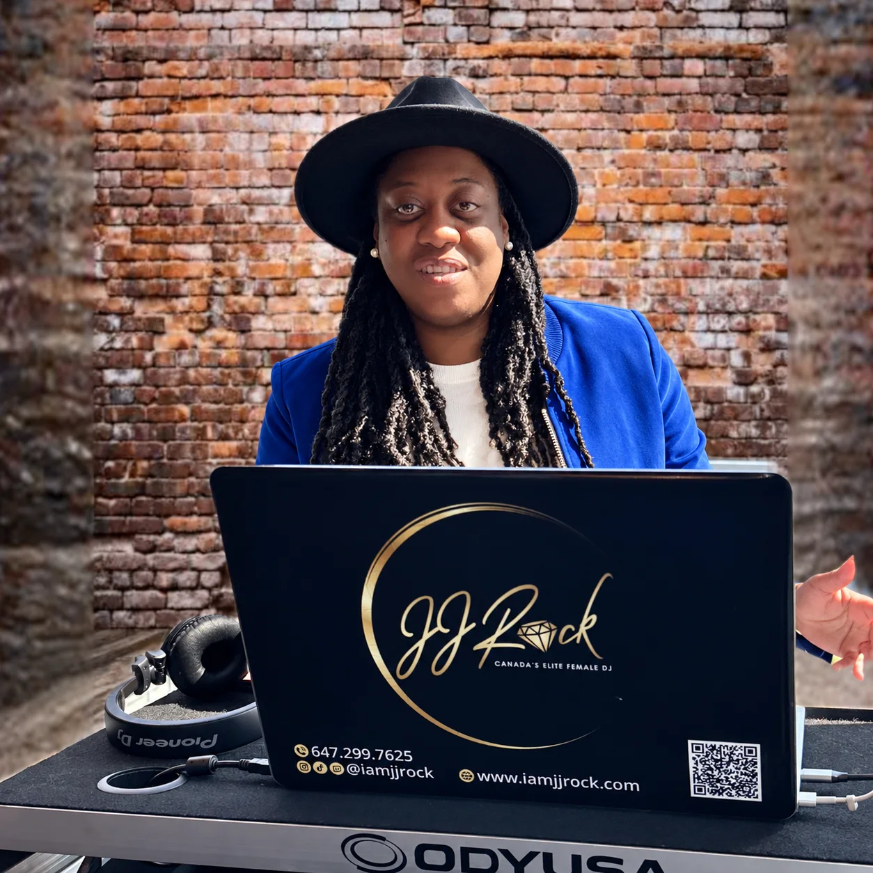 JJ Rock in a blue blazer and black hat behind her branded DJ laptop