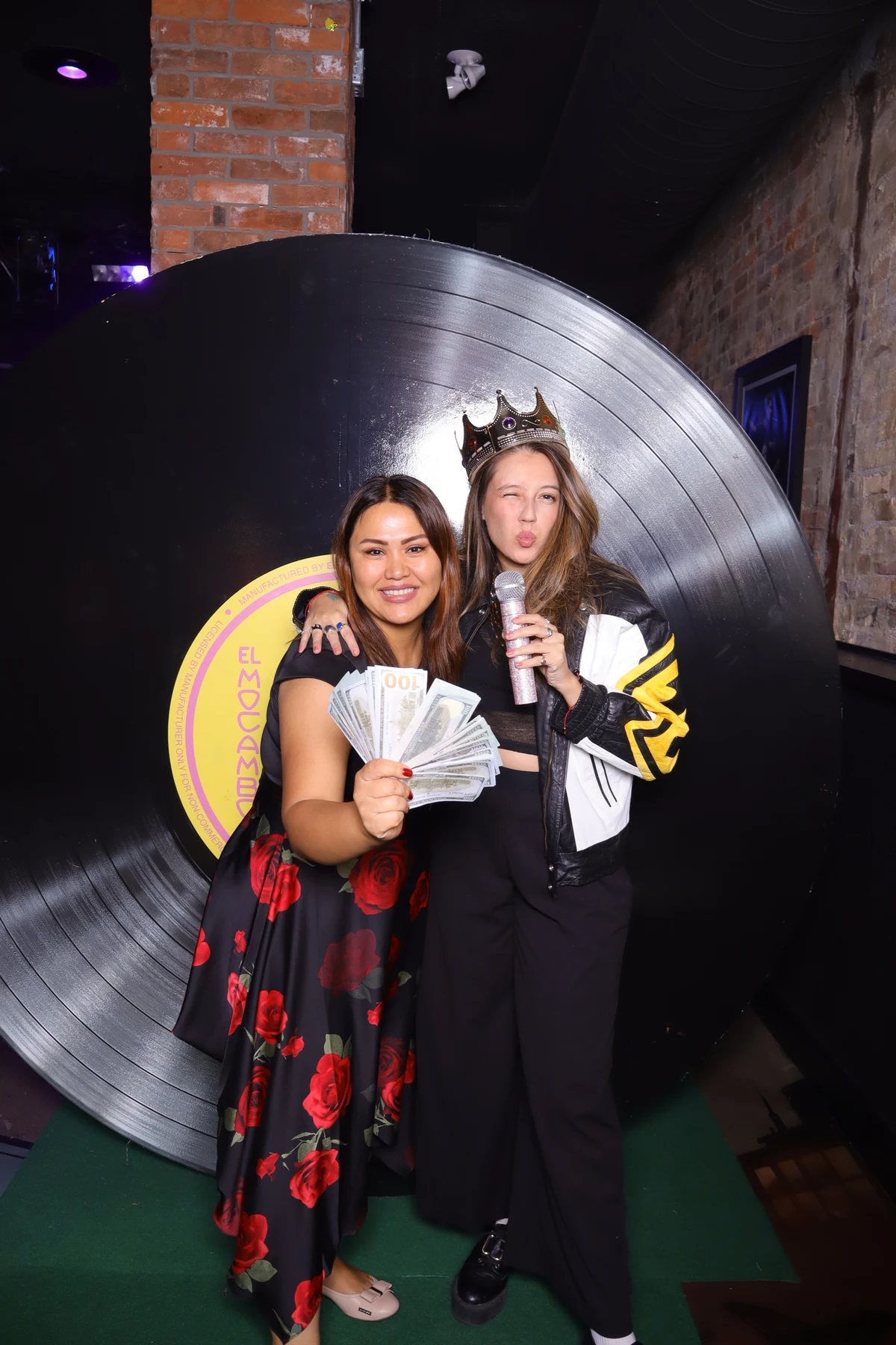 Two guests posing with a crown and microphone prop beside a record backdrop