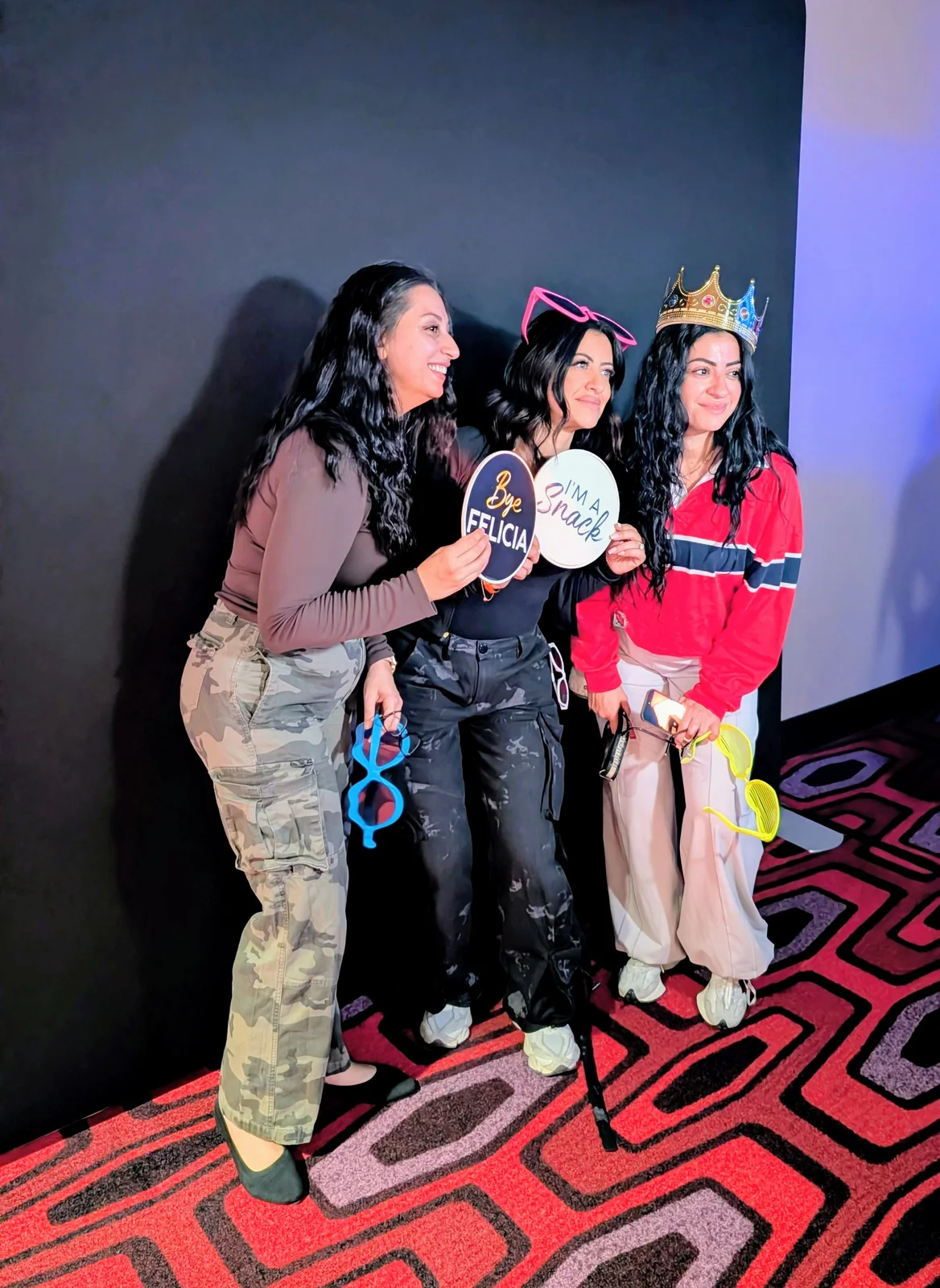 Three guests holding playful signs and wearing crown and glasses props against a black backdrop